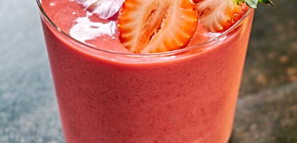 Pretty pink smoothie in a clear glass garnished with cut strawberries and a purple edible flower.